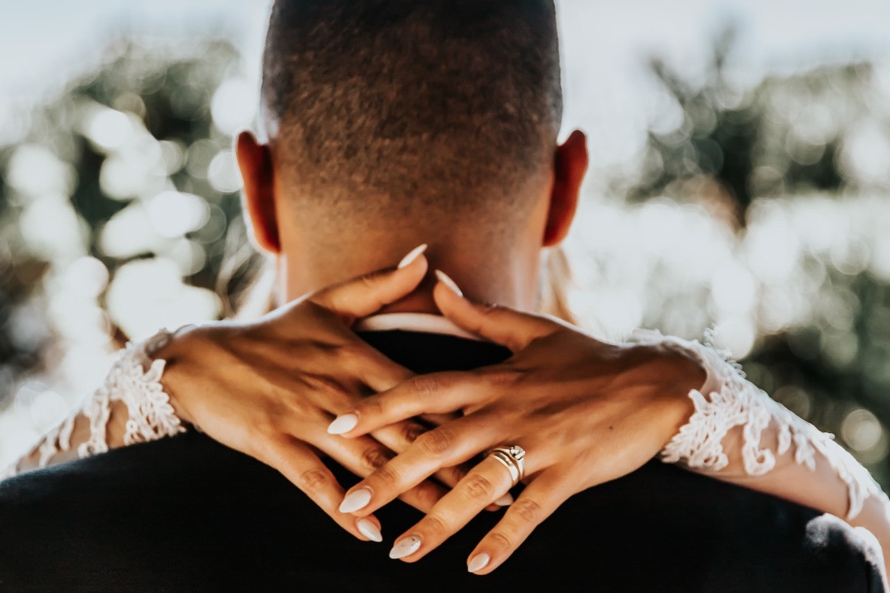 A husband and wife on their wedding day; the bride has her arms around his neck, showing her bridal set and nails along with edges of her detailed, long-sleeve gown.