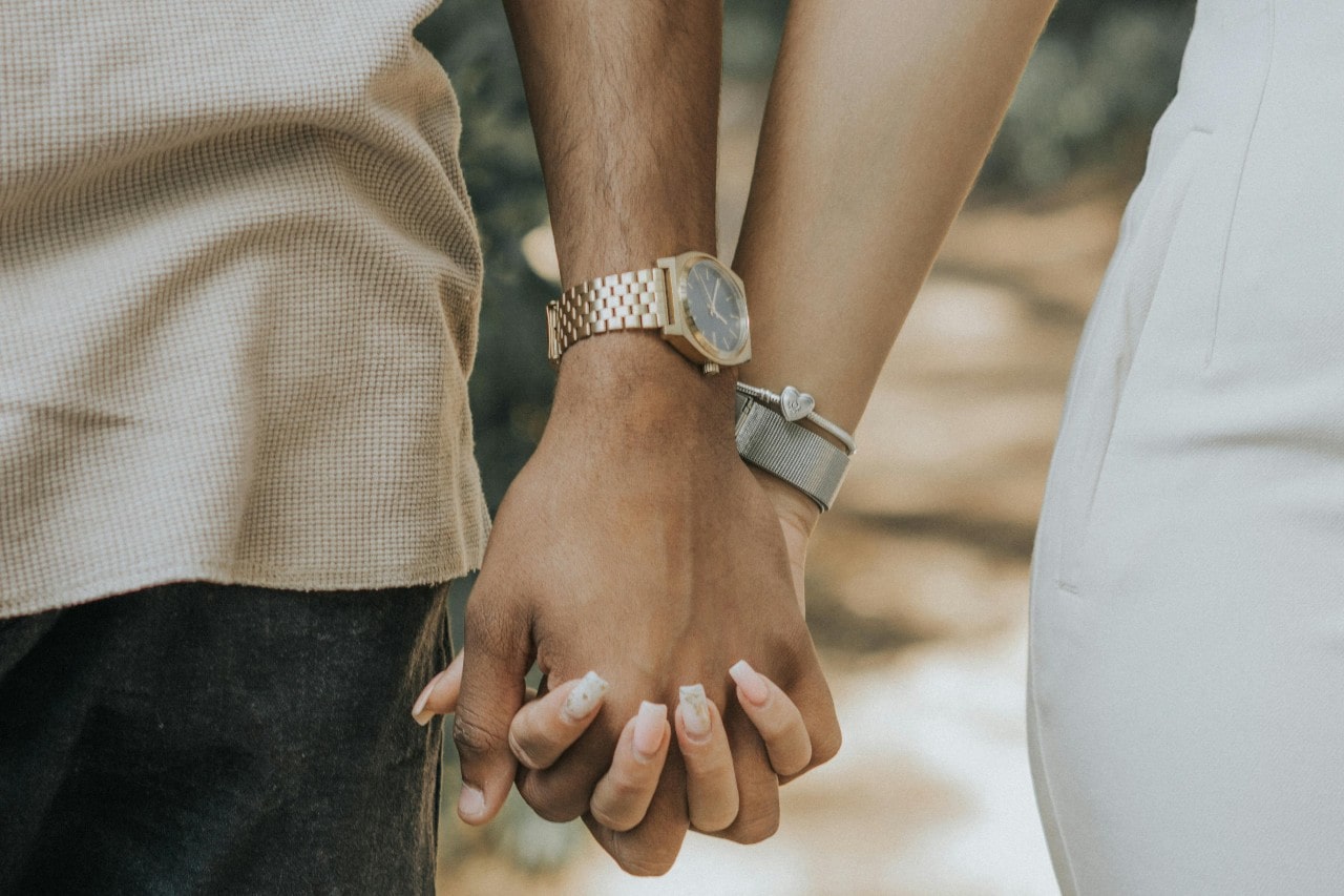 A couple holding hands, the man wearing a yellow gold watch and the woman wearing silver toned bracelets.