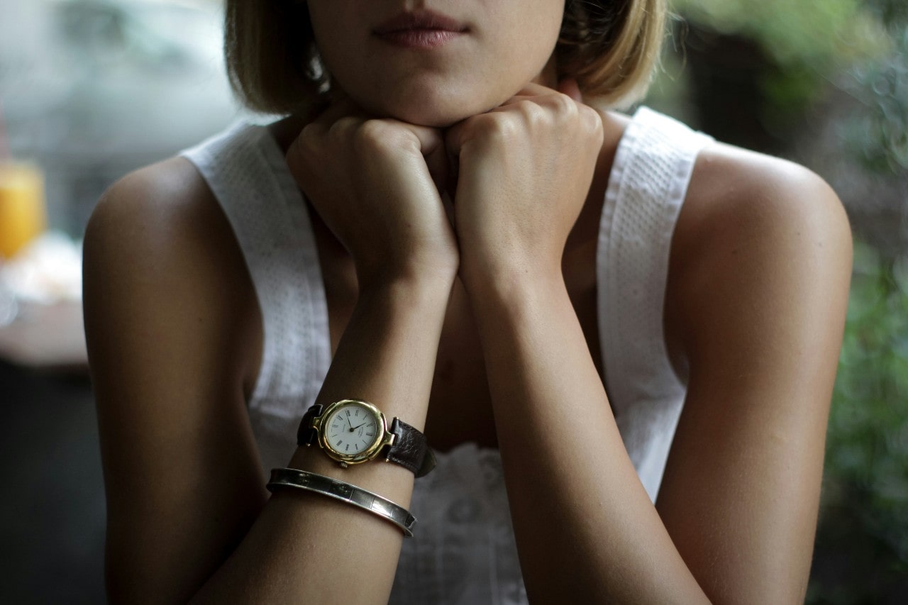 A woman resting her hands on her chin, wearing a yellow gold watch and silver bracelet.