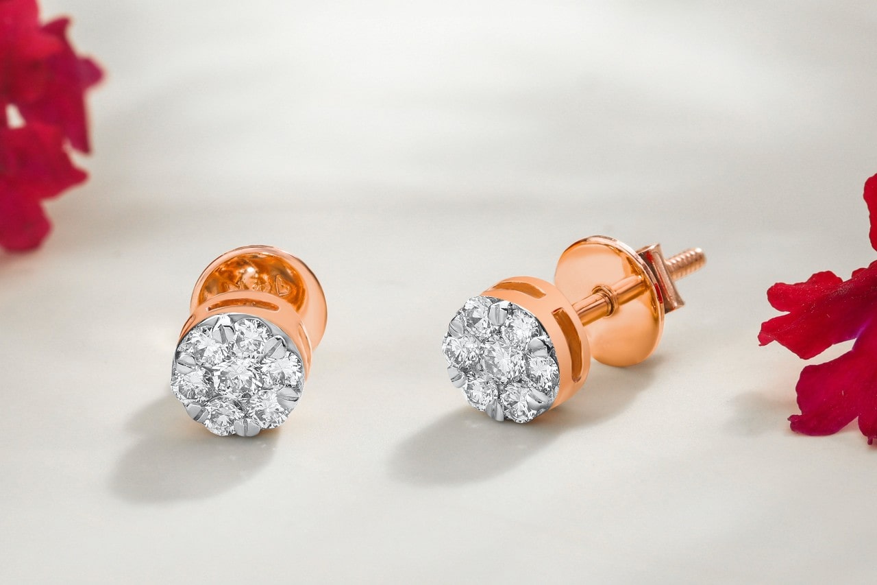 A pair of rose gold diamond halo stud earrings next to red flowers on a white surface.