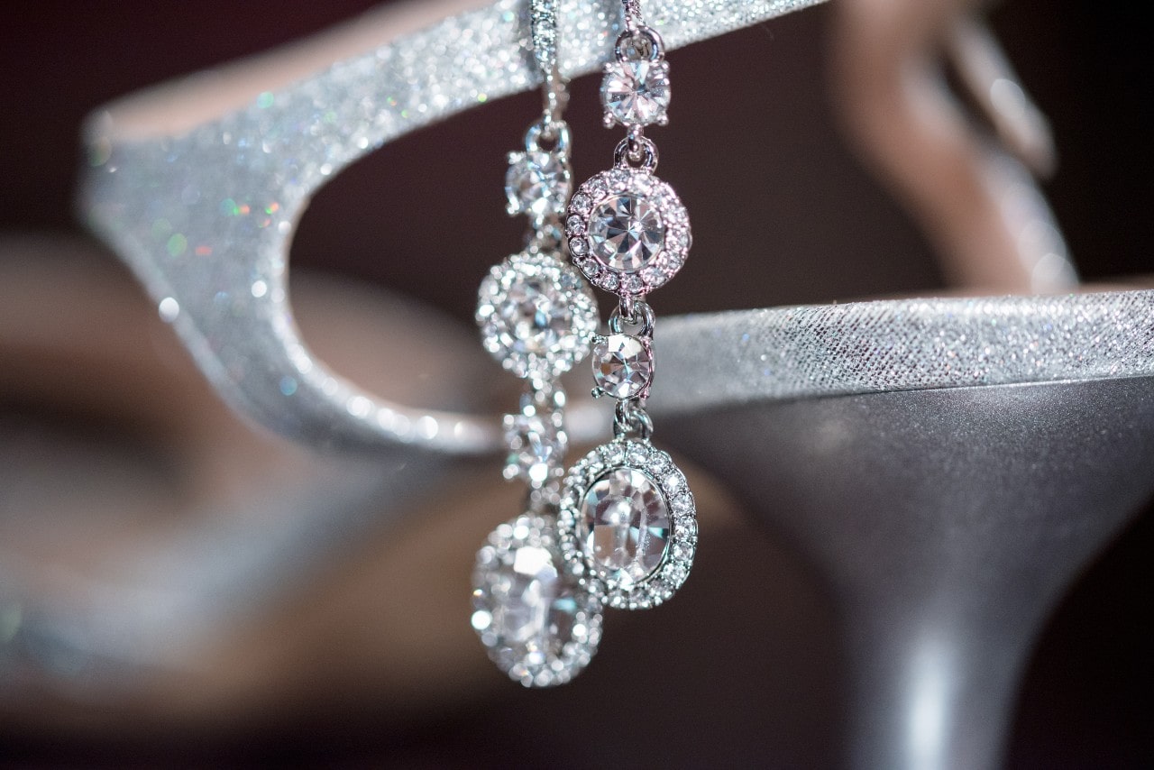 A pair of white gold diamond drop earrings hanging from a silver high heel.