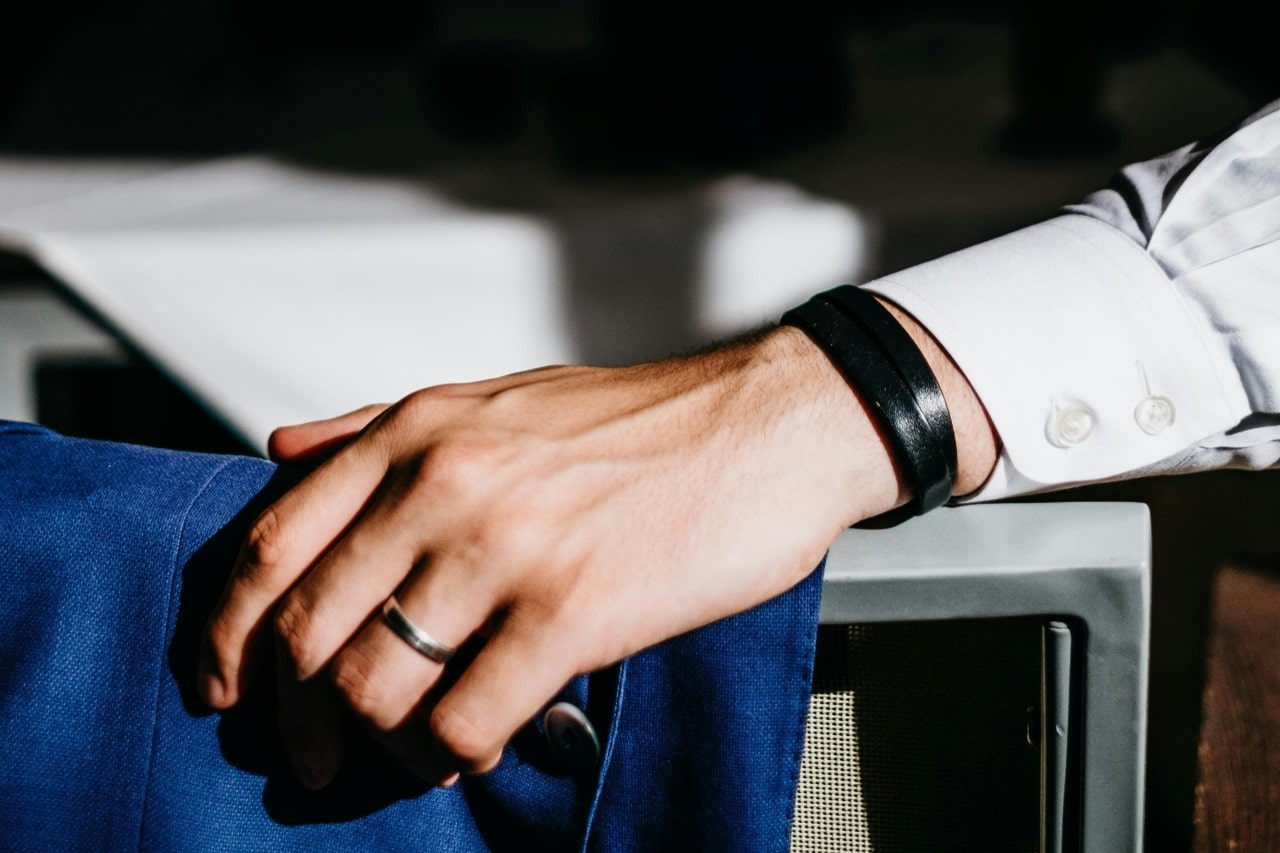 a man’s hand wearing a white gold wedding band resting on a chair back