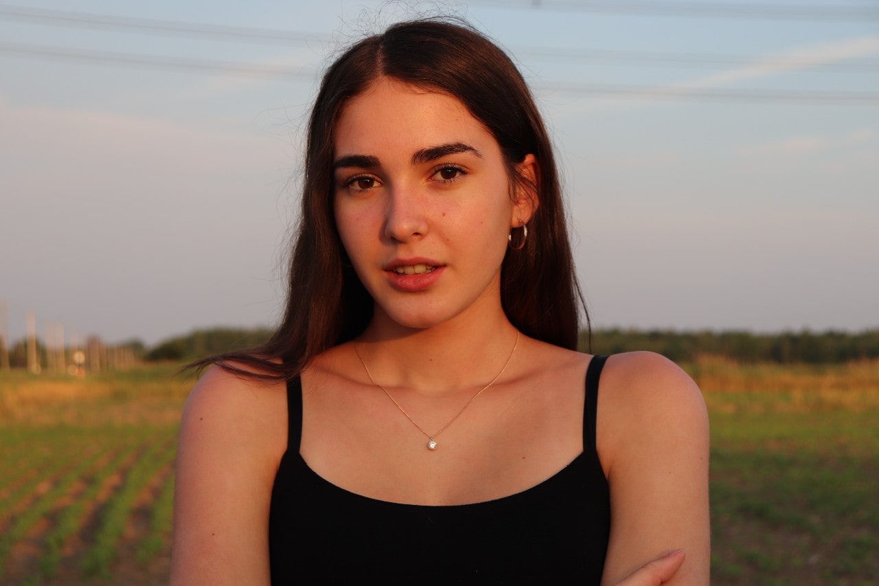 A close-up of a young woman standing in a field, wearing a simple diamond necklace.