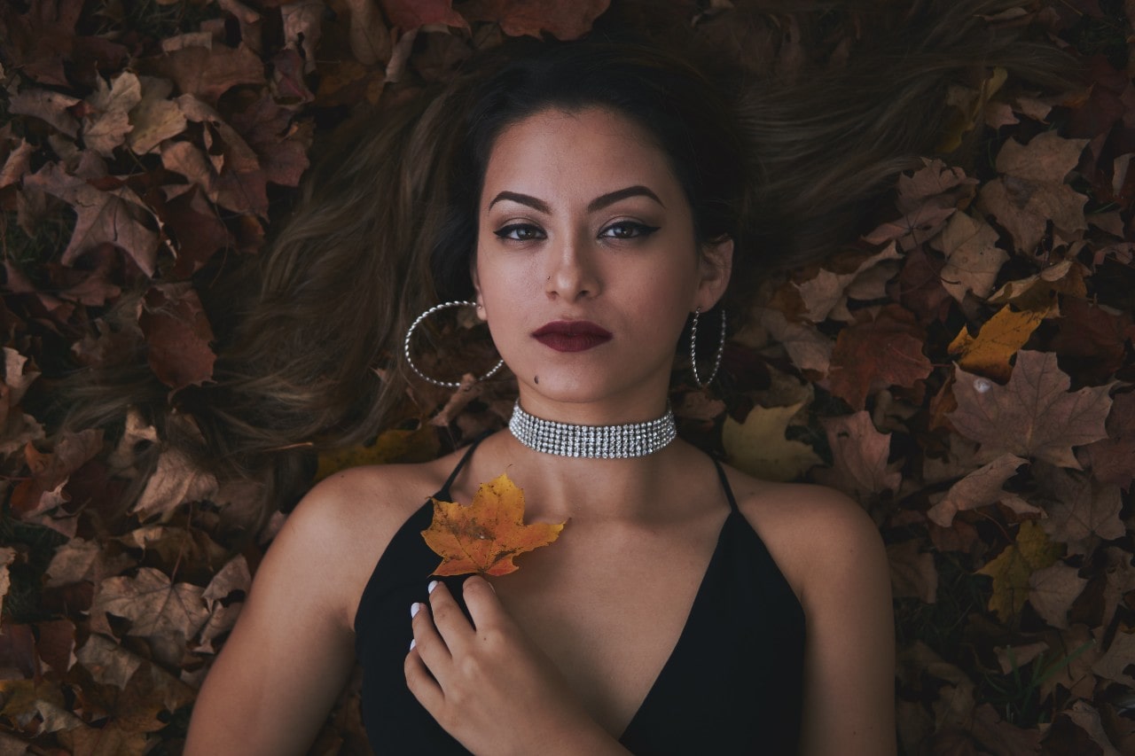 A close-up of an elegant woman laying in autumn leaves, wearing a bold diamond necklace and matching hoop earrings.