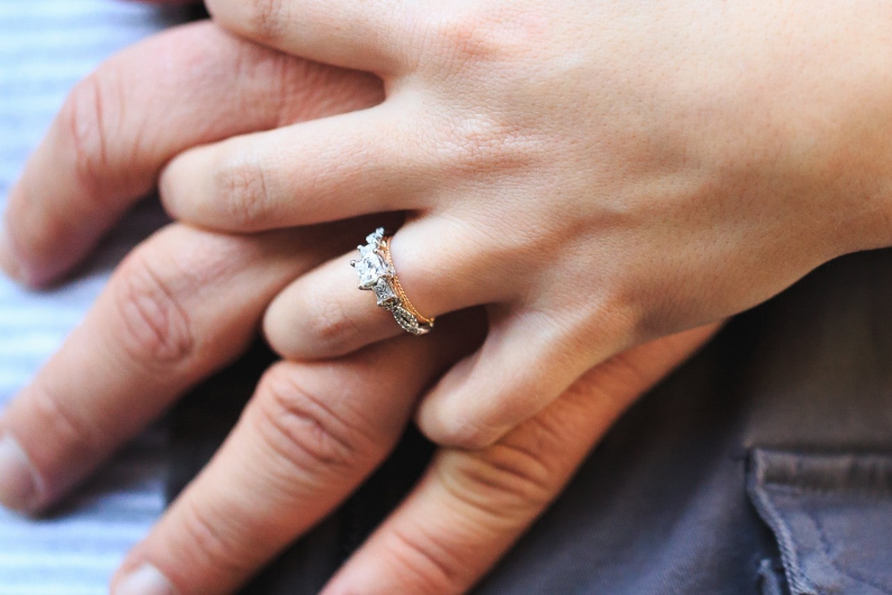 a pair of hands intertwined, the woman wearing a mixed metal three stone engagement ring