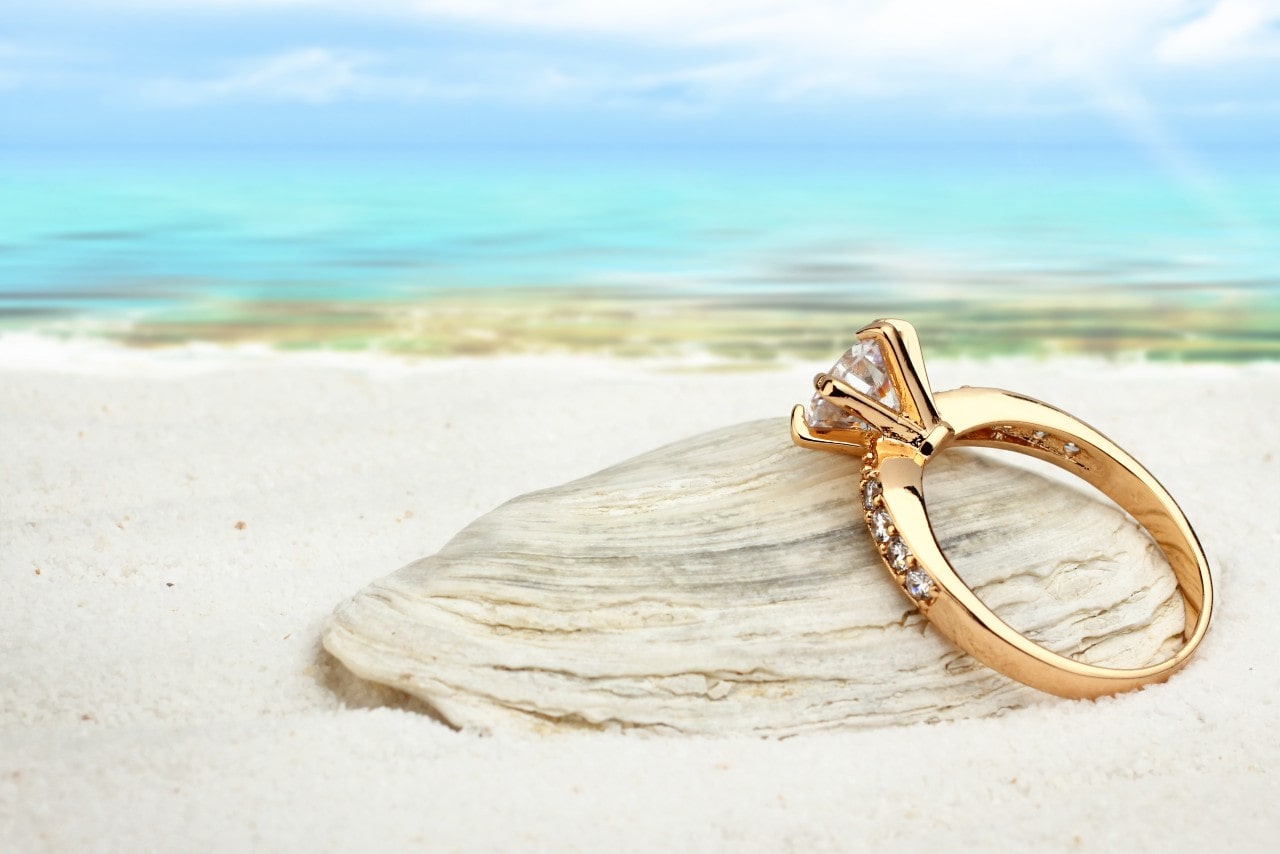 A close-up of an elegant engagement ring on a beach, resting on a clam shell.